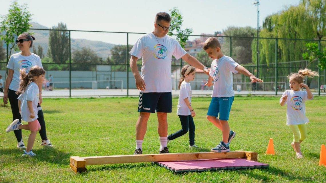 Kids guided by instructors at a park balance on a beam for fun and exercise.
