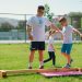 Kids guided by instructors at a park balance on a beam for fun and exercise.