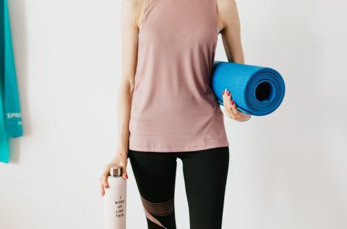 Faceless slim female athlete in sportswear standing with blue fitness mat and water bottle while preparing for indoors workout